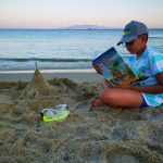 Zwycięzca konkursu. Chłopiec siedzi na plaży nad morzem, w czapce z daszkiem i błękitnej koszulce. Trzyma książkę. Przed nim zamek zbudowany z piachu.