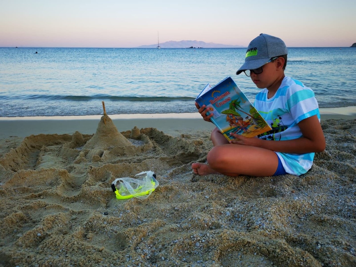 Zwycięzca konkursu. Chłopiec siedzi na plaży nad morzem, w czapce z daszkiem i błękitnej koszulce. Trzyma książkę. Przed nim zamek zbudowany z piachu.