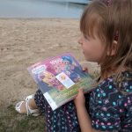 Mała dziewczynka ubrana w niebieską sukienkę w kwiatki siedzi na piachu nad brzegiem jeziora. Trzyma książkę.