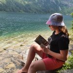 Dziewczynka czyta książkę. W tle jezioro górskie oraz góry.