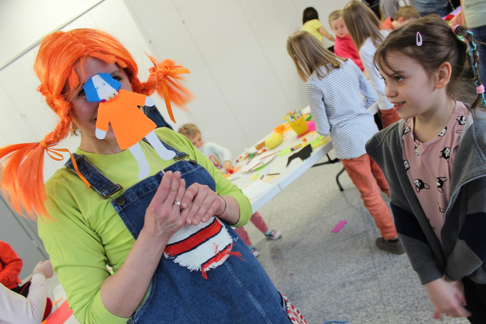 Kobieta w przebraniu Pippi Langstrumpf, ma na sobie kolorowy strój, pomarańczowe włosy, zakrywa twarz kukiełką . Obok stoi uśmiechnięta dziewczynka. W tle bawiące się dzieci.