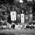 Czarno-biała fotografia przedstawia prawosławną procesję ze sztandarami, na pierwszym palnie wysoka trawa, w tle gęsta ścian wysokich drzew