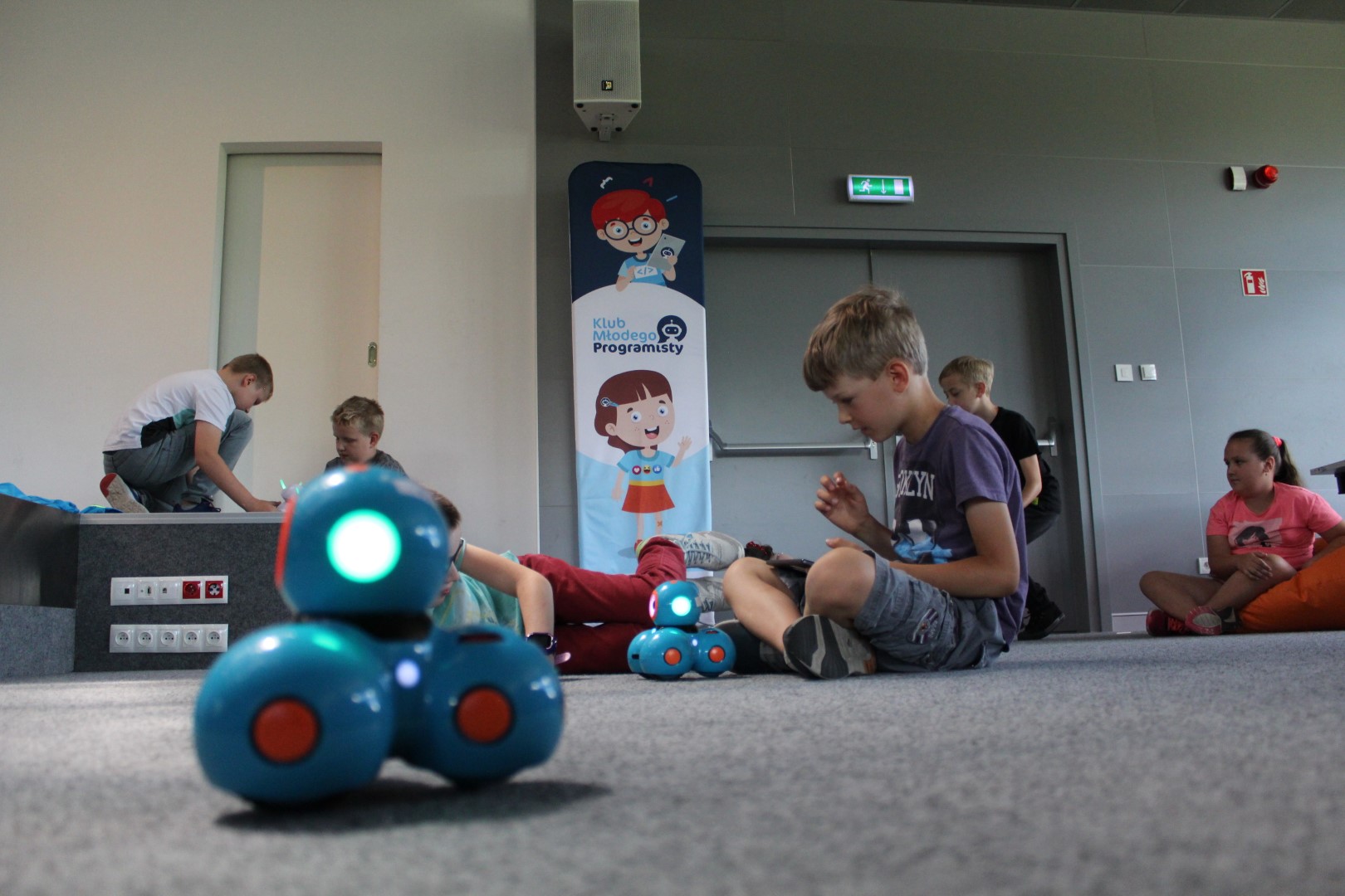 Na pierwszym planie robot, w tle grupa dzieci, która tworzy programy uruchamiające inne roboty.