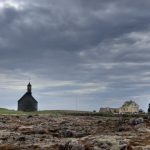 Islandia: na pierwszym tle skaliste podłoże z kamienną pryzmą, w tle sylwetka kościoła i zabudowań mieszkalnych. Niebo zachmurzone
