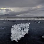 Islandia: Bryła lodu leży na czarnym piasku plaży, w tle morze, w oddali most i zarys lądu