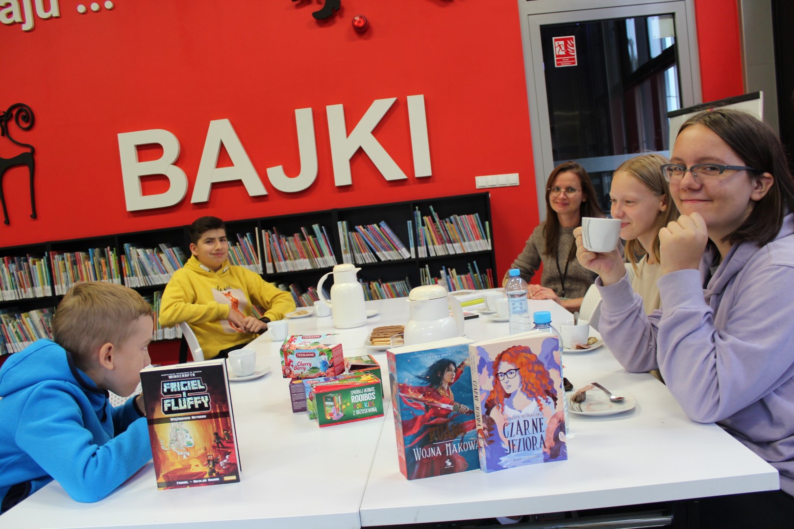 W pomieszczeniu Biblioteki wokół stołu siedzi czworo dzieci i bibliotekarka prowadząca spotkanie . Na stole są poustawiane filiżanki z talerzykami, herbata w pudełkach, dzbanki z wodą, ciastka na talerzu, książki. W tle na czerwonej ścianie - duży czerwony napis - bajki