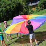 Słoneczny dzień. W otoczeniu zieleni na trawniku przed budynkiem Biblioteki grupa dzieci bawi się dużą okrągłą, kolorową chustą.
