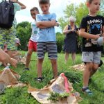 Słoneczny dzień. W otoczeniu zieleni na trawniku przed budynkiem Biblioteki grupa dzieci obserwuje erupcję stworzonych przez siebie wulkanów.