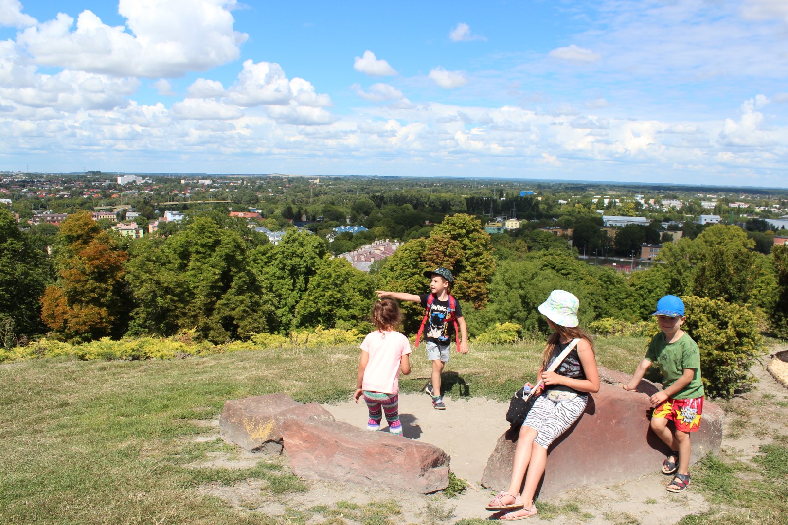 Słoneczny dzień. W otoczeniu zieleni, na górce chełmskiej grupa dzieci siedzi na kamieniach i obserwują otoczenie.