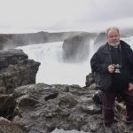 Mężczyzna z aparatem fotograficznym w dłoni pozuje na tle islandzkiego wodospadu