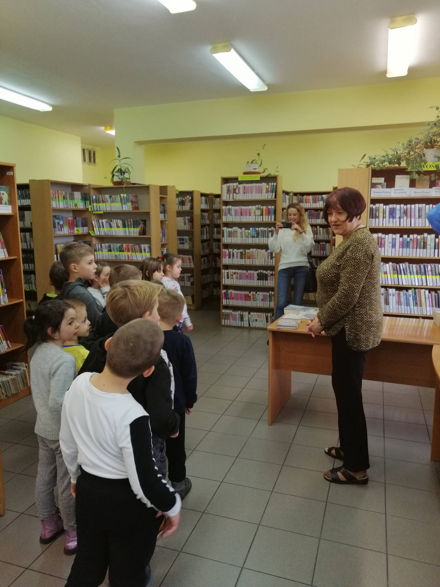 Pomieszczenie biblioteki: grupa dzieci jest witana przez bibliotekarkę