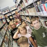 Grupa dzieci w wieku przedszkolnym stoi w Wypożyczalni Głównej wśród regałów z książkami