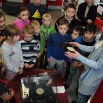 Grupa dzieci stoi w Wypożyczalni Zbiorów Audiowizualnych wokół gramofonu, który jest uruchamiany przez bibliotekarkę