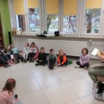 Dzieci siedzą w sali i słuchają opowiadania czytanego przez pracownika biblioteki