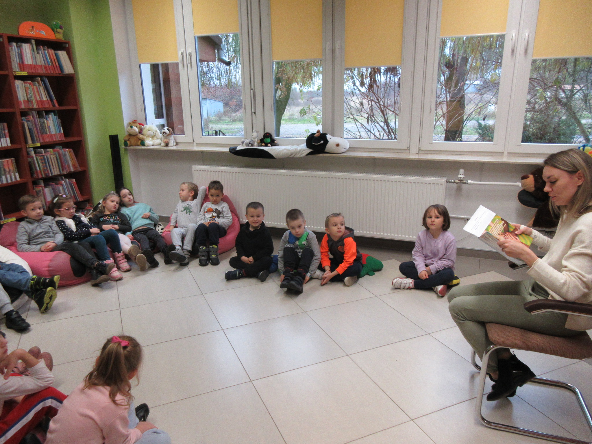 Dzieci siedzą w sali i słuchają opowiadania czytanego przez pracownika biblioteki