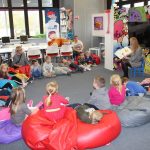 Grupa dzieci w wieku przedszkolnym siedzi w okręgu w Wypożyczalni dla dzieci, słuchają czytanej przez bibliotekarkę książki