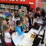 Grupa dzieci w wieku przedszkolnym stoi w Wypożyczalni dla dzieci wokół gramofonu, który jest uruchamiany przez bibliotekarkę