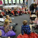 Wypożyczalni dla dzieci: Bibliotekarka rozdaje dzieciom siedzącym w kręgu na podłodze upominki z drewnianej skrzyni