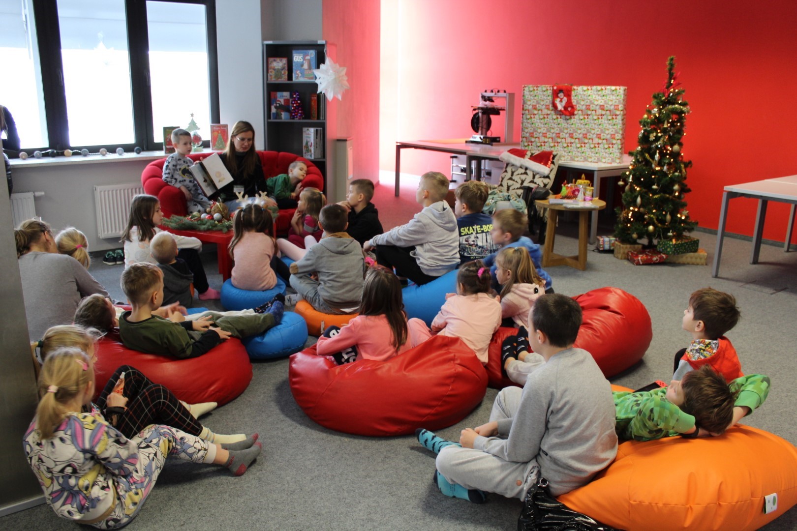 W pomieszczeniu Biblioteki grupa dzieci siedzi na podłodze w kolorowych pufach, zwróceni są do kobiety , która czyta książkę