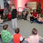 W pomieszczeniu biblioteki dzieci wraz z kobietą siedzą na podłodze tworząc koło. W środku stoi chłopiec i się rozgląda