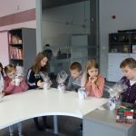W pomieszczeniu biblioteki dzieci stoją wokół stołu i oglądają pomalowane kubki