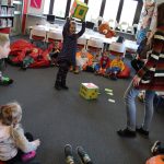 Wypożyczalnia dla dzieci biblioteki . Dzieci siedzą wokół Pani bibliotekarki. Na środku stoi chłopiec, który rzuca kostką