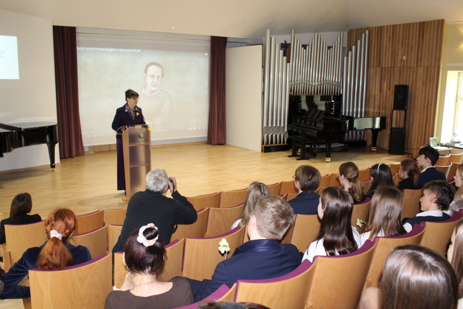 Na scenie sali koncertowej Państwowej Szkoły Muzycznej stoi prelegentka która wygłasza wykład z okazji 80 rocznicy powstania w getcie warszawskim