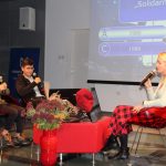 Uczestnicy turnieju wiedzy z okresu lat osiemdziesiątych w sali widowiskowe biblioteki wraz z prowadzącą siedzą przed ekranem komputera. W tle ekran z wyświetlonymi pytaniami