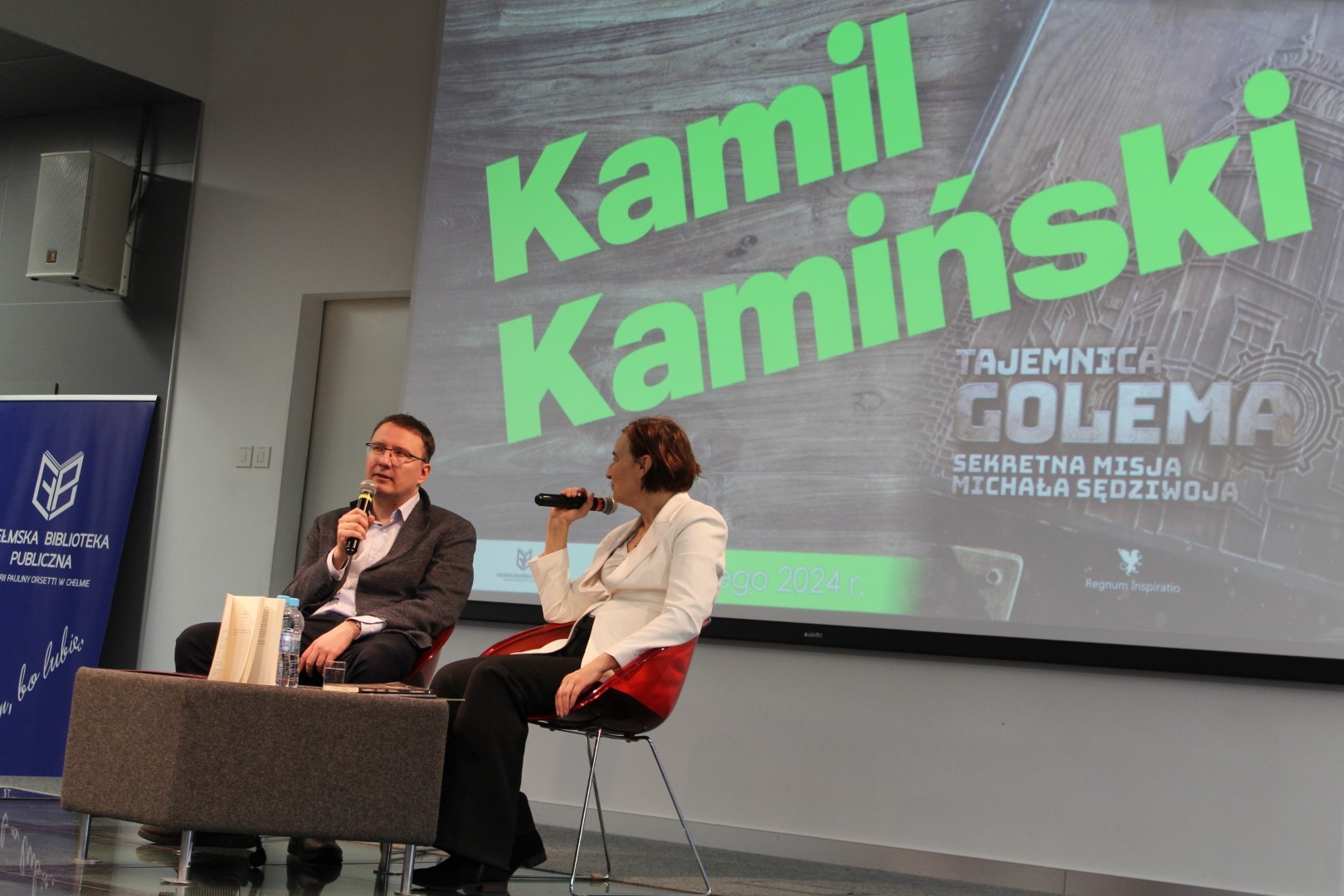 Spotkanie autorskie z Kamilem Kamińskim. Na scenie autor z prowadzącą na drugim planie ekran z zielonym napisem