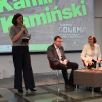 Spotkanie autorskie z Kamilem Kamińskim. Na scenie przy stoliki siedzi autor z prowadzącą, obok stoi kobieta, która przemawia, na drugim planie ekran z zielonym napisem