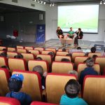 Dwóch mężczyzn i kobieta siedzą na krzesłach na scenie sali widowiskowej biblioteki