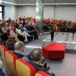 Na scenie sali widowiskowej biblioteki kobieta w tle widzowie