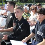 Publiczność siedzi w otoczeniu drzew w pełnym słońcu i patrzy przed siebie