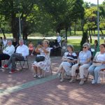 Publiczność siedzi w otoczeniu drzew w pełnym słońcu i patrzy przed siebie