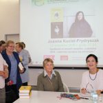 Uczestnicy spotkania wraz z pisarką pozują do zdjęcia. W tle ekran z plakatem