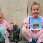 Dwie dziewczynki patrzą w obiektyw. Uśmiechają się. Jedna trzyma książkę