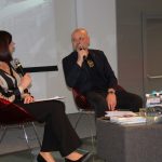 Na scenie sali widowiskowej dwie osoby siedzą na krzesłach i przemawiają do publiczności