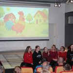 Grupa uczennic siedzi na scenie sali widowiskowej i śpiewa. w tle animacja z kogutem