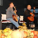 Dwaj muzycy graja na gitarach, na pierwszym planie dynie i jesienne liście