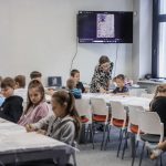 Uczestnicy warsztatów z kaligrafii siedzą przy stolikach. Na drugim planie prowadząca zajęcia