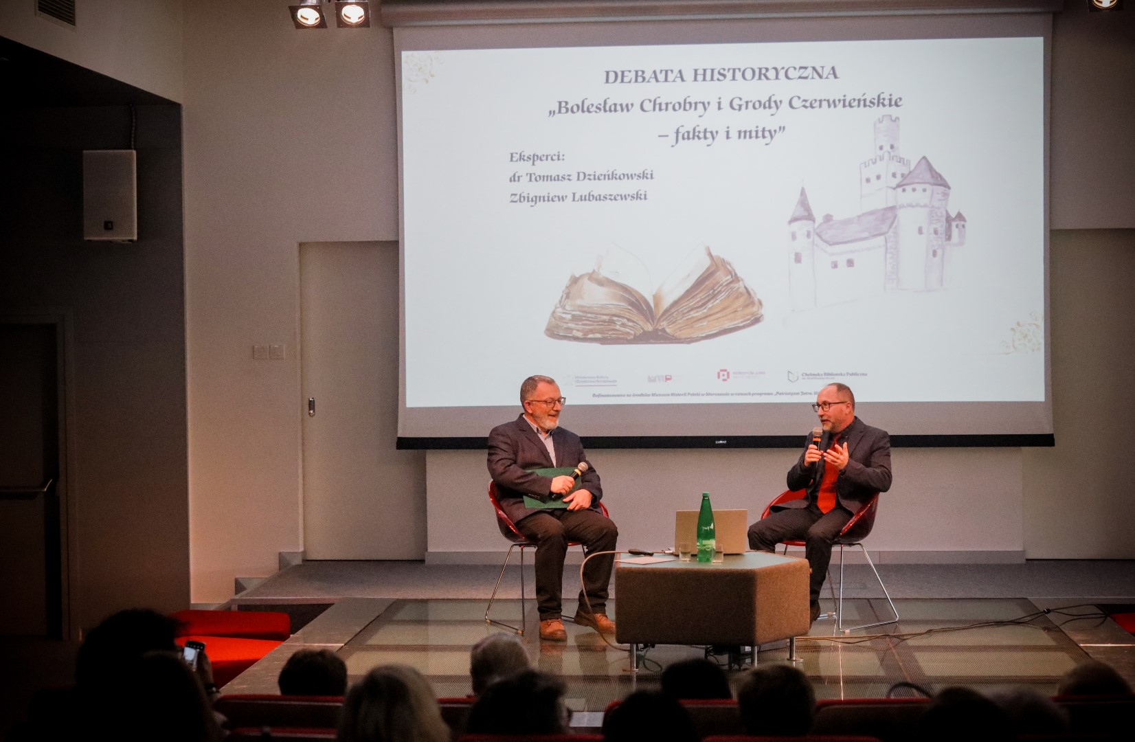 Mężczyźni siedzą na scenie sali widowiskowej. W tle fragment przygotowanej prezenatcji
