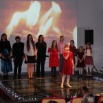 Dzieci stoją na scenie i śpiewają w tle ekran z płomieniami