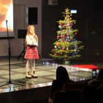 Dziewczynka stoi na scenie w tle choinka i ekran z płomieniami