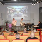 Grupa osób na scenie sali widowiskowej czytają poezję